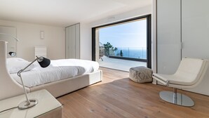 Un dormitorio moderno con cama, lámpara de noche, silla y un gran ventanal con vista al mar.