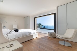 A modern bedroom with a bed, bedside lamp, chair, and a large window offering a view of the sea.