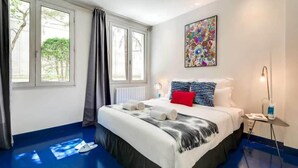 A hotel room with a large bed, a bedside table with a lamp, a colorful abstract painting on the wall, and a view of trees through the window. - Modern & colourful 3BD for 9 guests in Le Marais! (Paris)