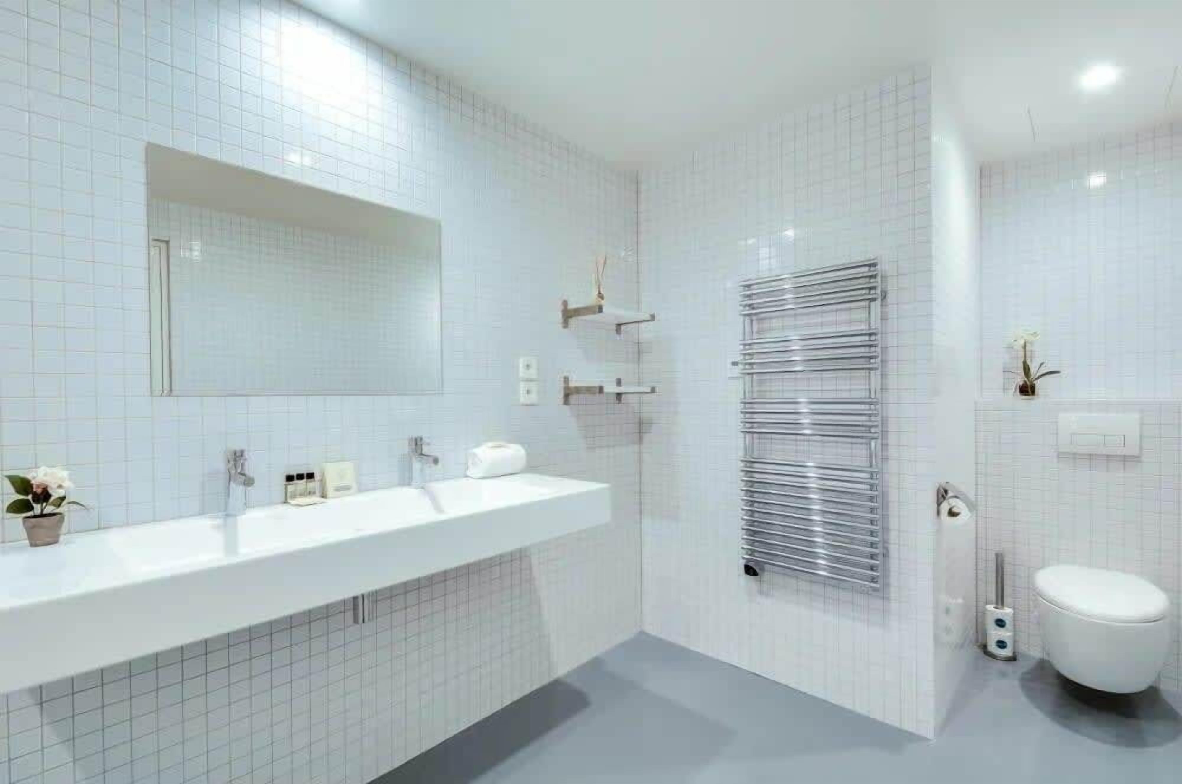 A modern bathroom with a white tiled wall, a large mirror, a white sink, a towel rack, and a toilet.