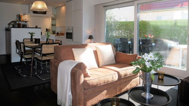 A modern living room with a sofa, dining table, and chairs, a kitchen area, and a large window with a view. - Isabelle A1 - Bright family apartment with sunny terrace - in the concession (De Haan)