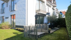 A two-story building with a brick facade, a balcony, and a small garden area with a wooden fence. - Isabelle A1 - Bright family apartment with sunny terrace - in the concession (De Haan)
