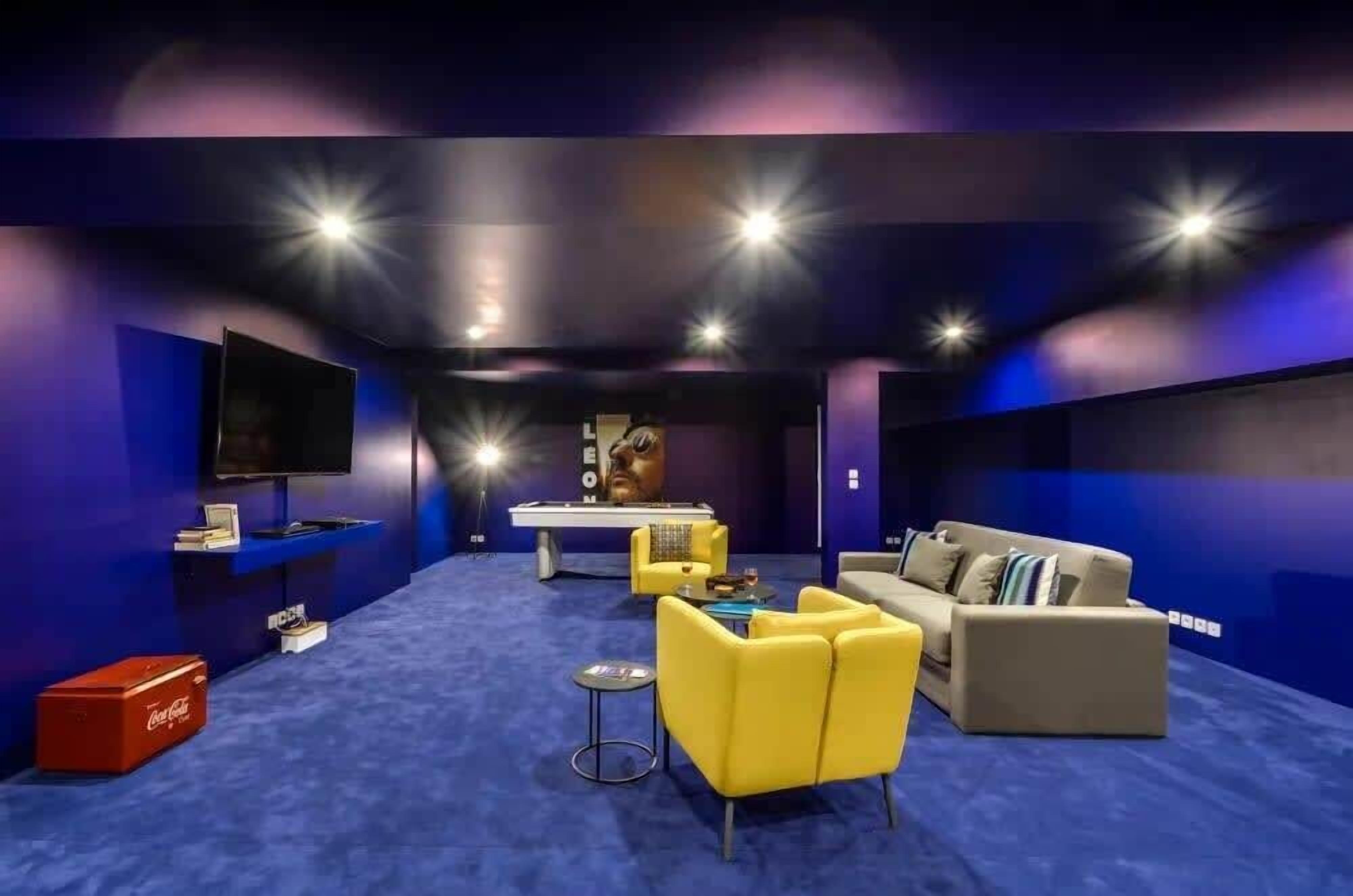 Una sala de cine moderna con alfombra azul, mobiliario amarillo y gris, una pantalla grande y una bola de discoteca.