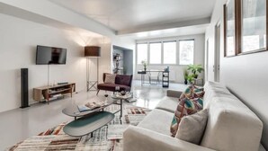 Ein modernes Wohnzimmer mit Sofa, Couchtisch und Fernseher. Es gibt Fenster, eine Stehlampe und ein Bücherregal.