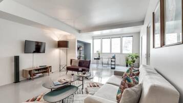 A modern living room with a sofa, coffee table, and TV. There are windows, a floor lamp, and a bookshelf.