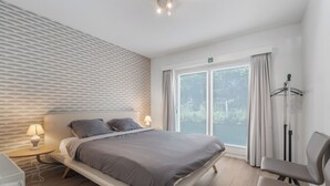 A modern hotel room with a large bed, bedside tables, a chair, and a view of the outdoors through a large window.