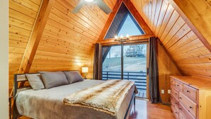 A cozy bedroom with a wooden sloped ceiling, a bed with a fur throw, a dresser, and a large window with a view of a house and trees.
