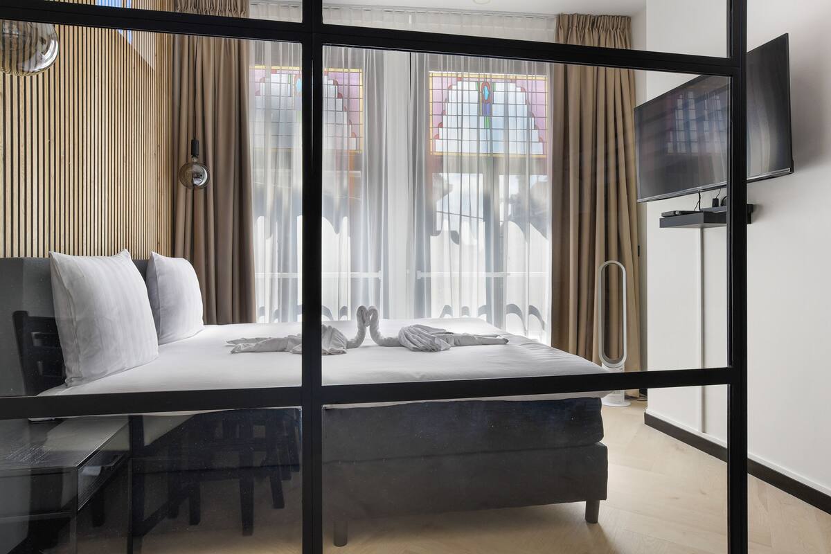 A modern hotel room with a bed, a desk, a chair, a TV, and a large window with curtains.