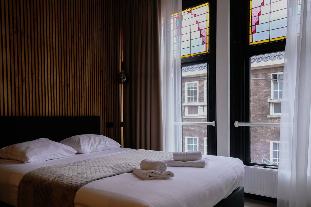 A neatly made bed with white linens and a folded towel, a wooden slatted headboard, and a view of a brick building through large windows with stained glass.