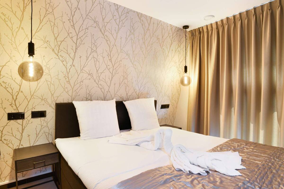 A hotel room with a bed, two bedside tables, a wall with a tree branch pattern, and a window with curtains.