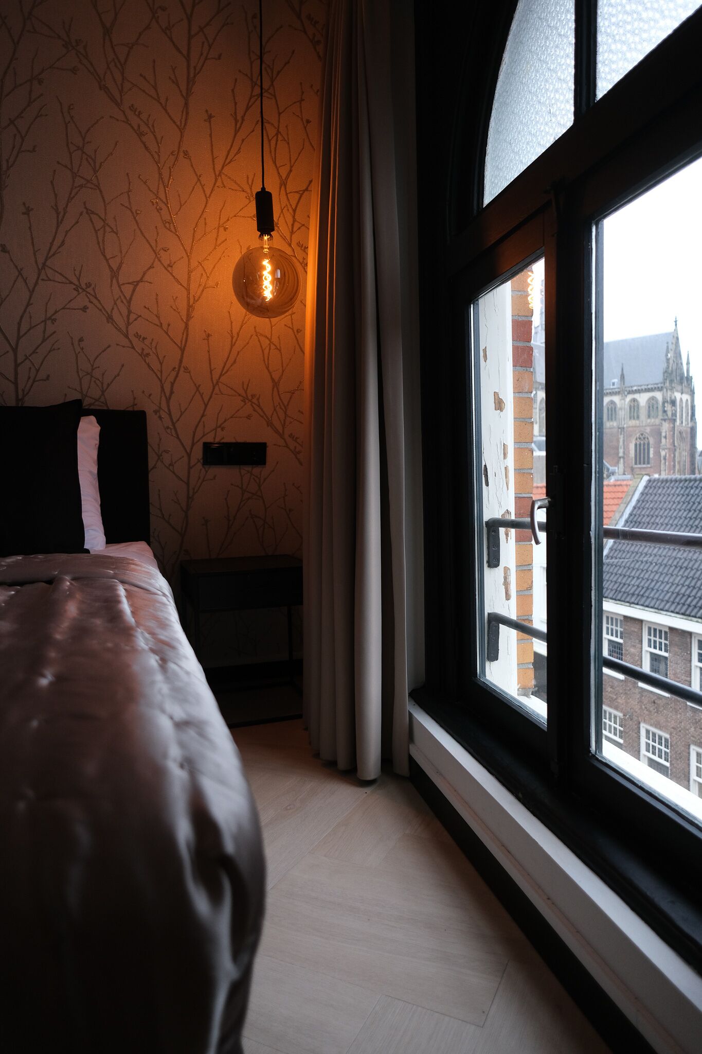A bedroom with a large bed, a hanging light fixture, a window with a view of buildings, and a curtain.
