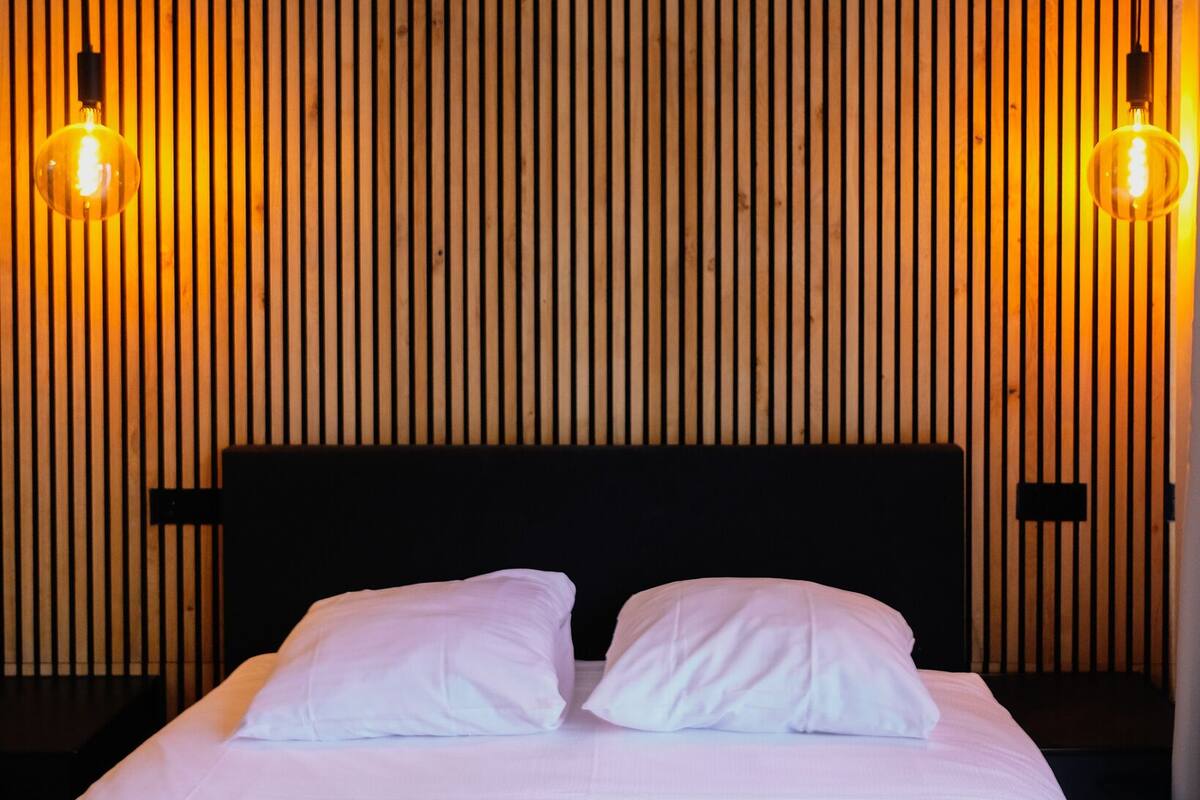 A bed with white bedding, two pillows, and a headboard against a wooden slat wall with two wall-mounted lights.