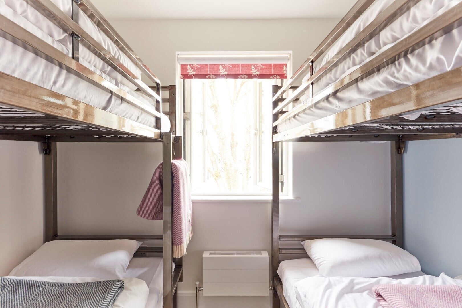 A dormitory room with bunk beds, a window with a red curtain, and a radiator.