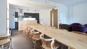 A modern kitchen with dark cabinets and a central island, a long wooden table with chairs, and a small round table with stools. - The Barrel Store is a unique hostel in the heart of the Cotswolds. (CIRENCESTER)