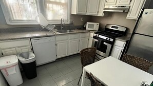 A compact kitchen with white cabinets, a microwave, stove, and a refrigerator. There is a small dining table with chairs and a broom leaning against the wall. - Clover2900 Apartment & Rooms in S Philly (Philadelphia)