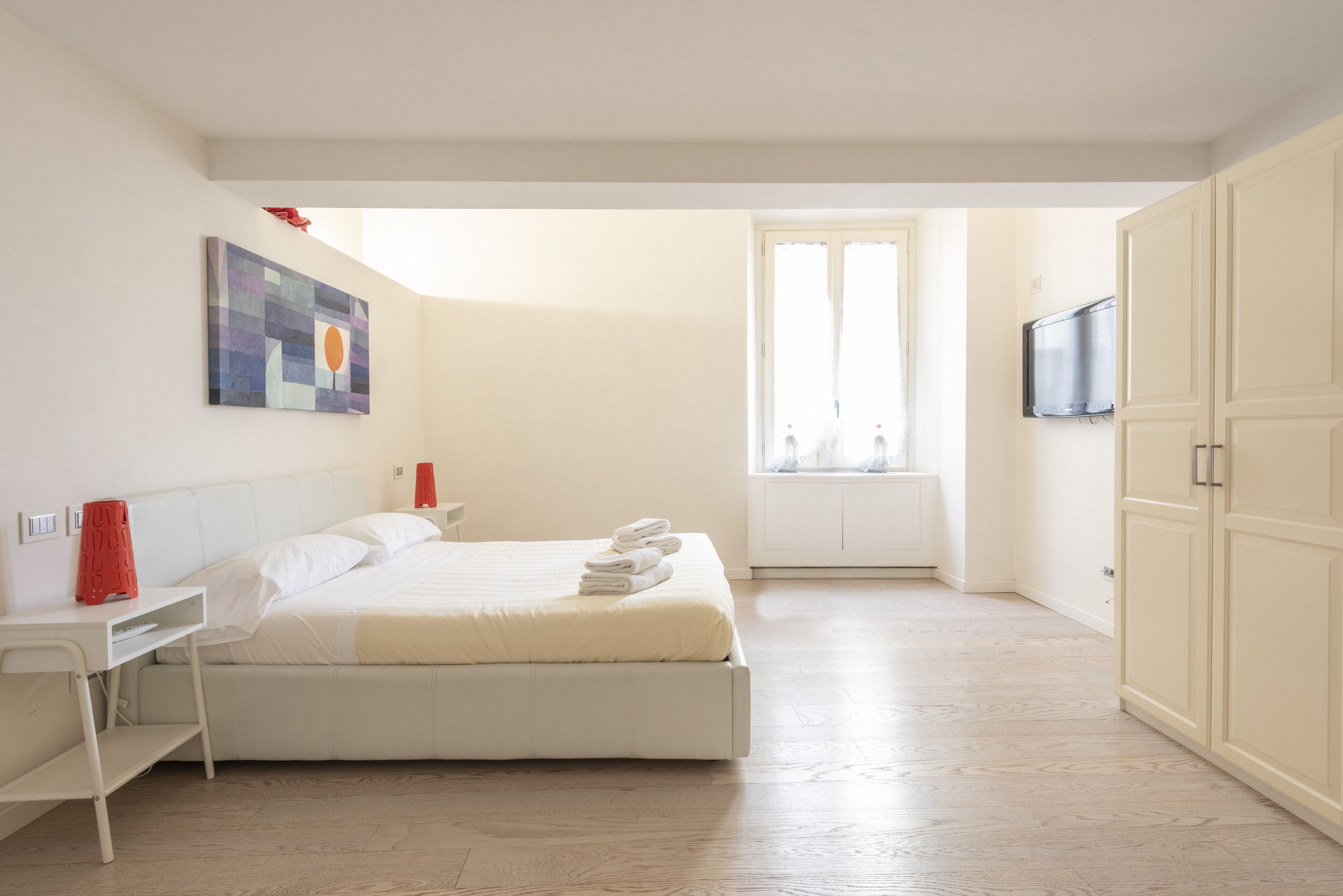 A bedroom with a bed, a desk, a chair, a TV, and a window.