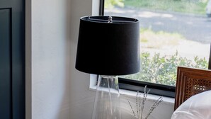A neatly made bed with a white pillow and a striped blanket, a small wooden nightstand with a black lamp, a white vase with dried flowers, and a book titled "Ellen DeGeneres."