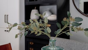 A white table with a glass vase containing green foliage. Behind the table, there is a black cabinet with glass doors, and a round mirror on the wall.
