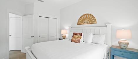 A bedroom with a large bed, a ceiling fan, a wall-mounted decorative piece, two bedside tables with lamps, and a door leading to another room.