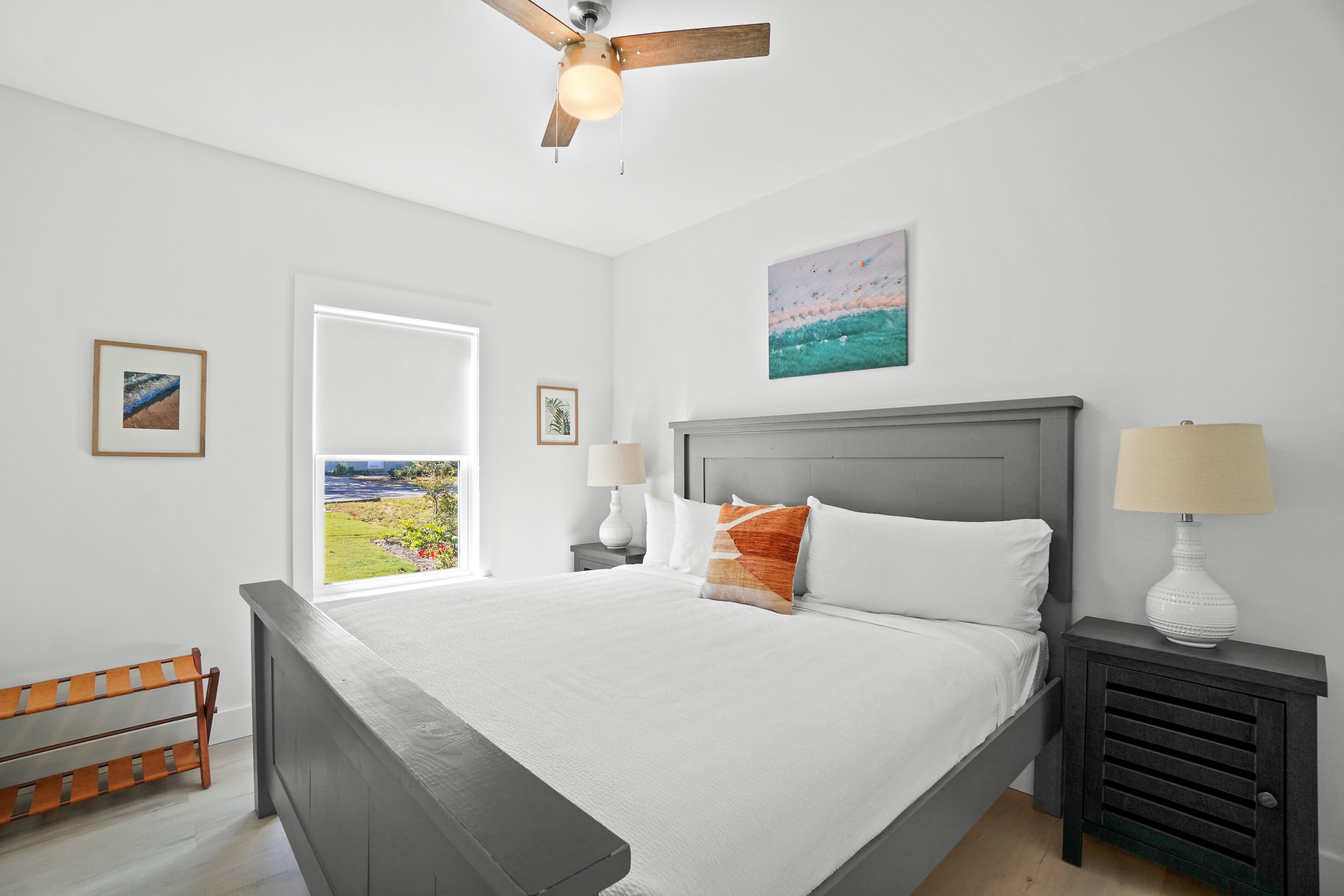 A bedroom with a large bed, a ceiling fan, a nightstand with a lamp, a bench, and a window with a view of a garden.