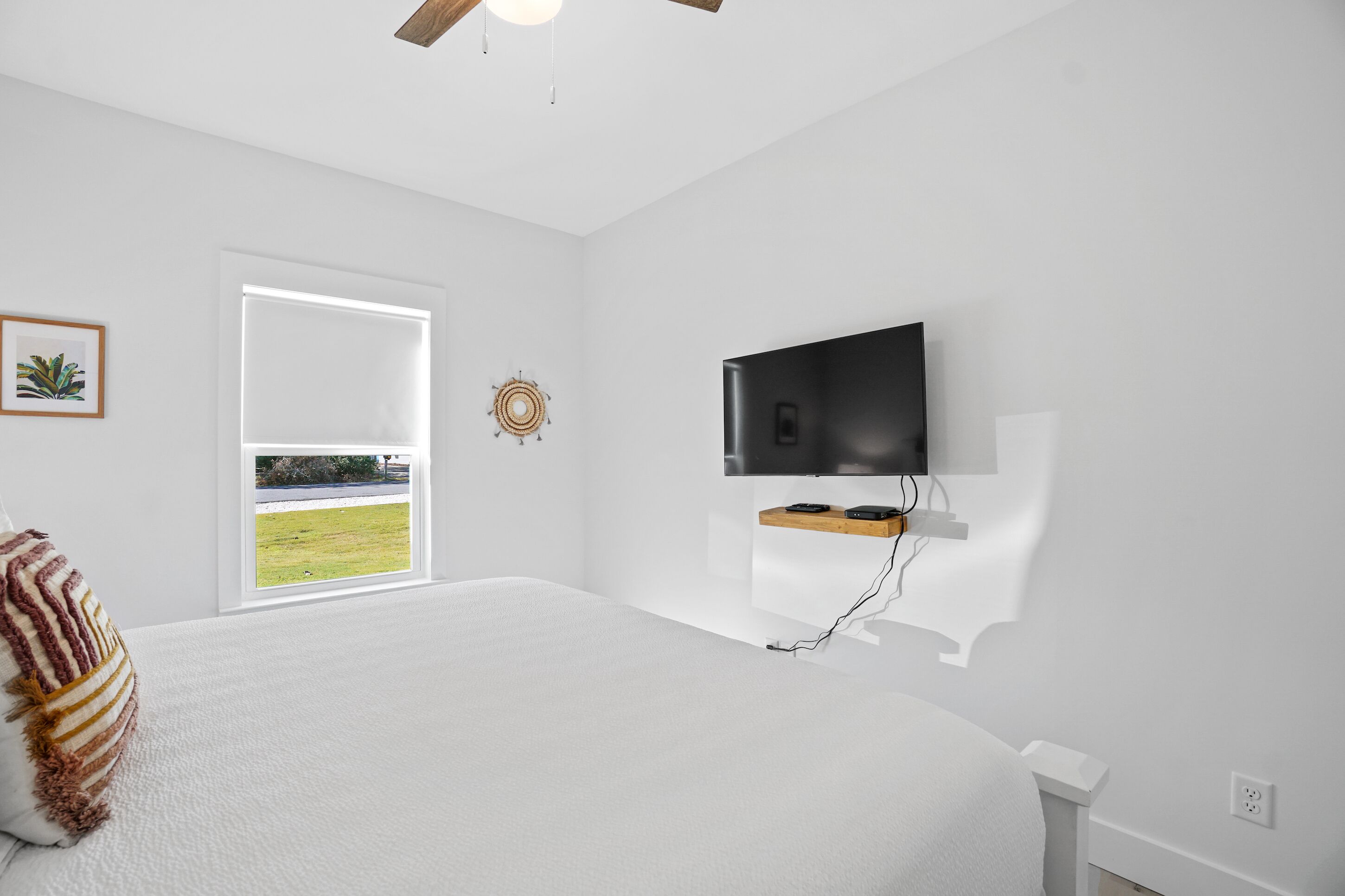 Un dormitorio con una cama grande, televisor montado, ventana con vista a un área verde y ventilador de techo.