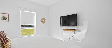 Un dormitorio con una cama grande, televisor montado, ventana con vista a un área verde y ventilador de techo.