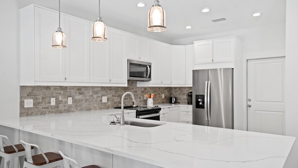 A modern kitchen with white cabinets, a large island with bar stools, stainless steel appliances, and pendant lighting. - Beach House - The Sanderling by PHG (Panama City Beach)