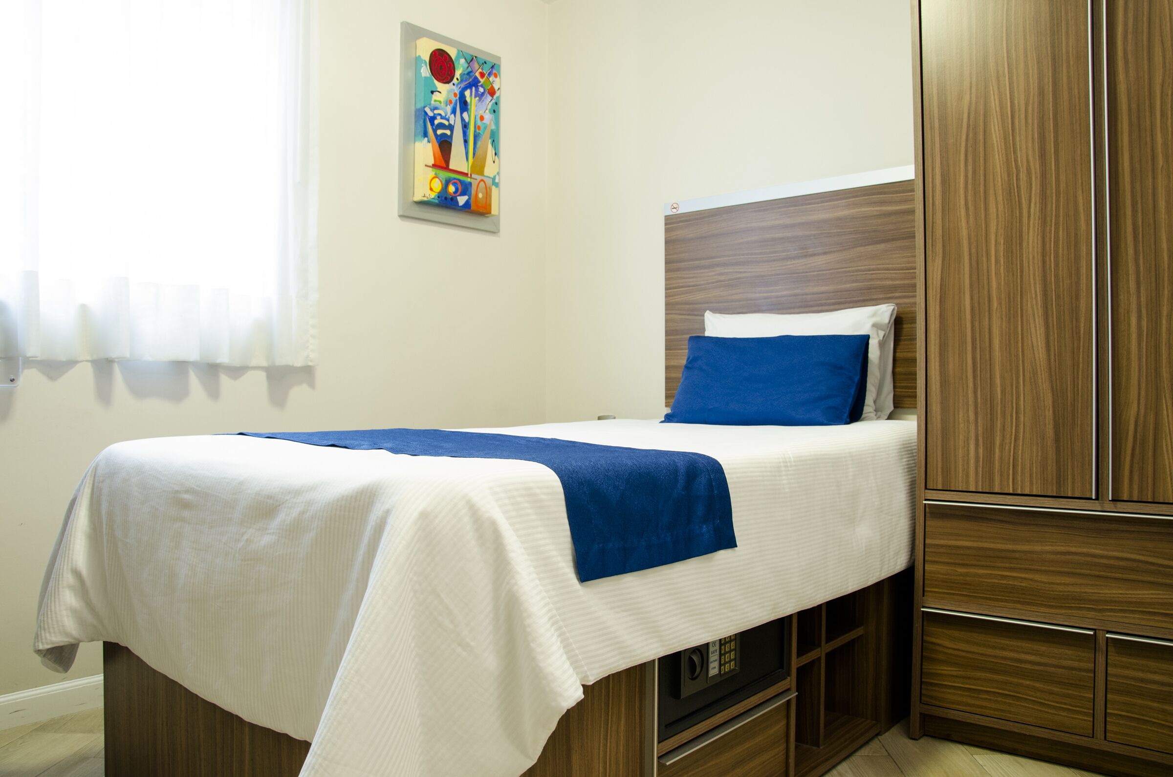 A neatly made bed with a blue pillow and white linens, a wooden wardrobe, and a colorful abstract painting on the wall.
