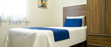 A neatly made bed with a blue pillow and white linens, a wooden wardrobe, and a colorful abstract painting on the wall.