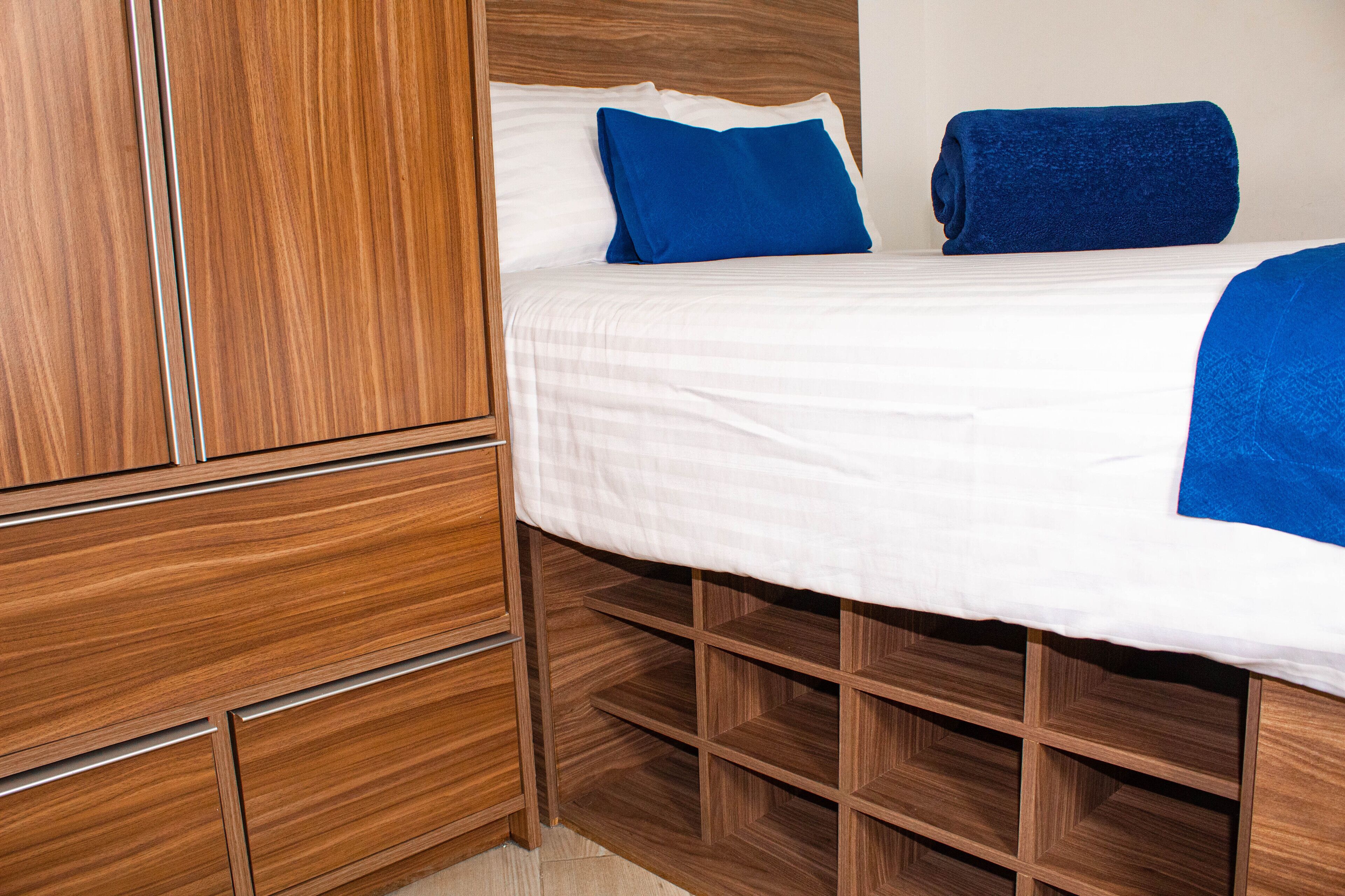 Ein ordentlich bezogenes Bett mit weißen Leinentüchern und blauen Kissen, ein Holzschrank mit mehreren Fächern sowie ein Holz-Kopfteil.
