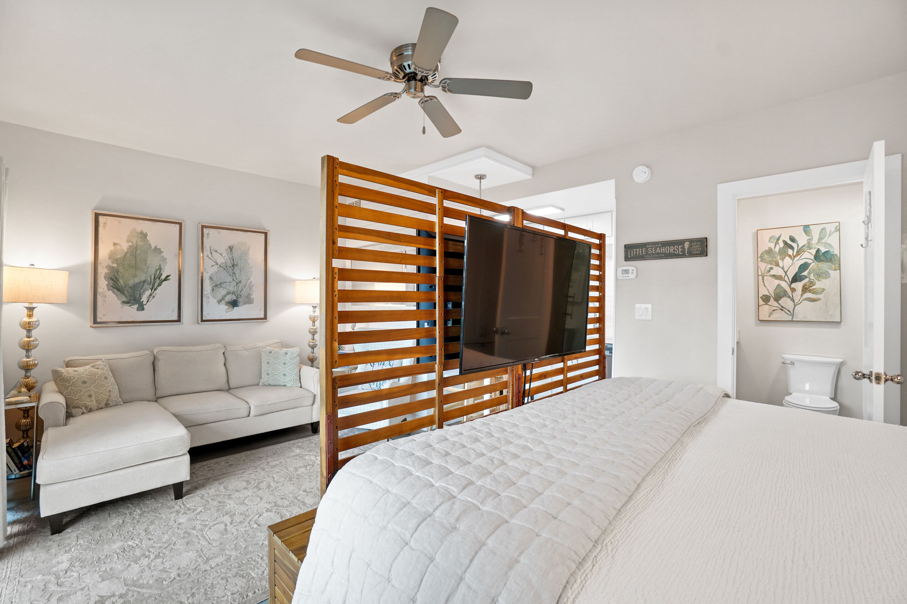 A bedroom with a bed, a sofa, a TV mounted on a wooden partition, a ceiling fan, and artwork on the walls.
