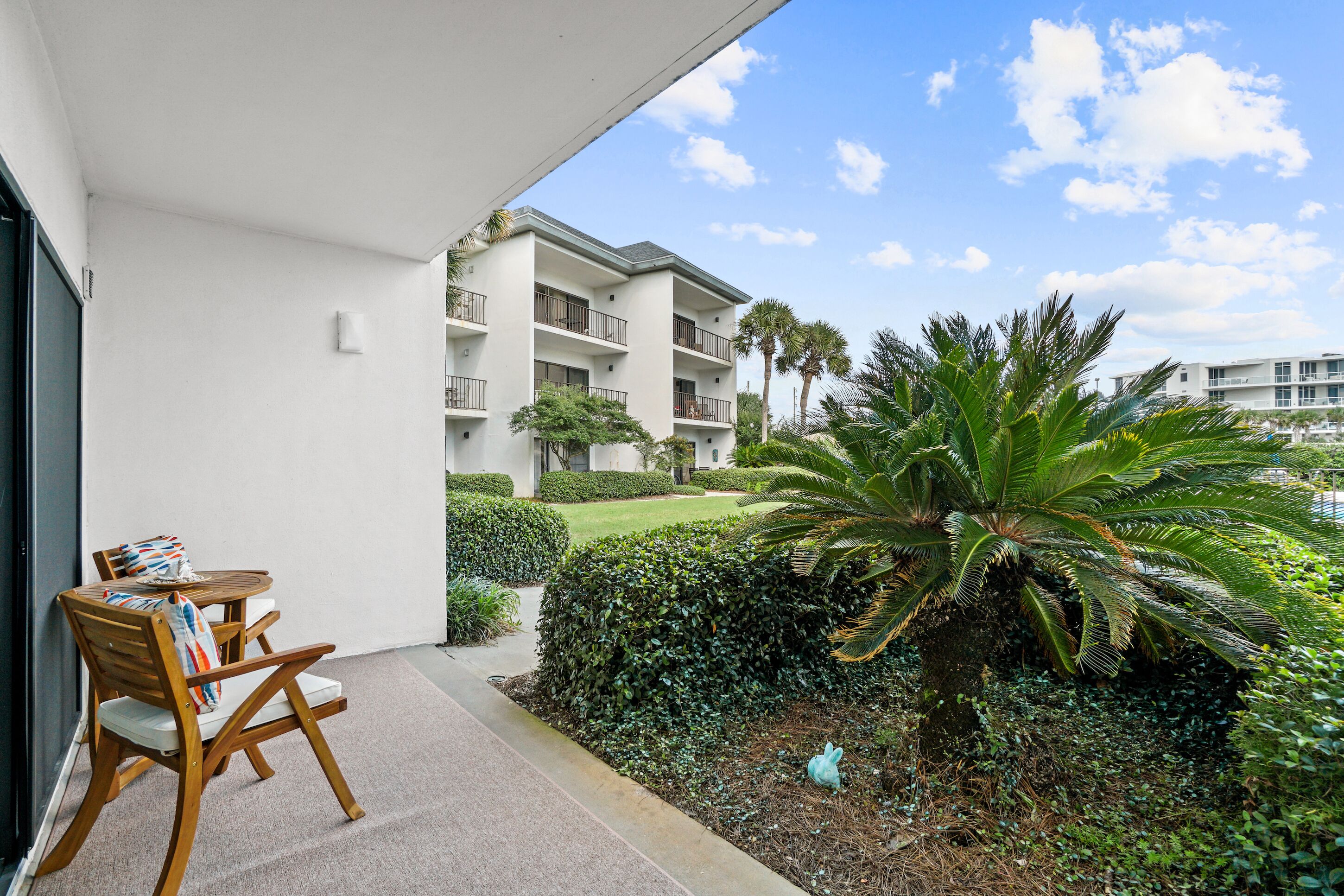 A balcony with a wooden chair and table, a white building in the background, and a well-maintained garden with a large palm tree.