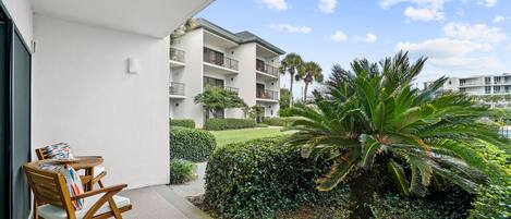 A balcony with a wooden chair and table, a white building in the background, and a well-maintained garden with a large palm tree.