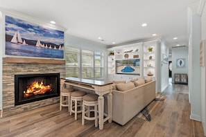 A modern living room with a fireplace, a bar area, and a large window overlooking a scenic view. - Pearls by the Sea - Luxury Home in Atlantic Beach (North Myrtle Beach)