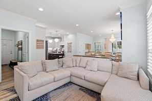 A modern living room with a large L-shaped sectional sofa, a kitchen in the background, and a dining area with wooden furniture. - Pearls by the Sea - Luxury Home in Atlantic Beach (North Myrtle Beach)
