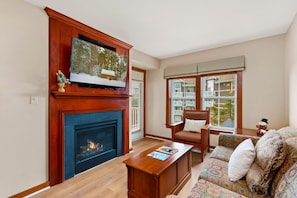 A living room with a fireplace, a television mounted on a wooden mantel, a sofa with patterned cushions, and a window with blinds.
