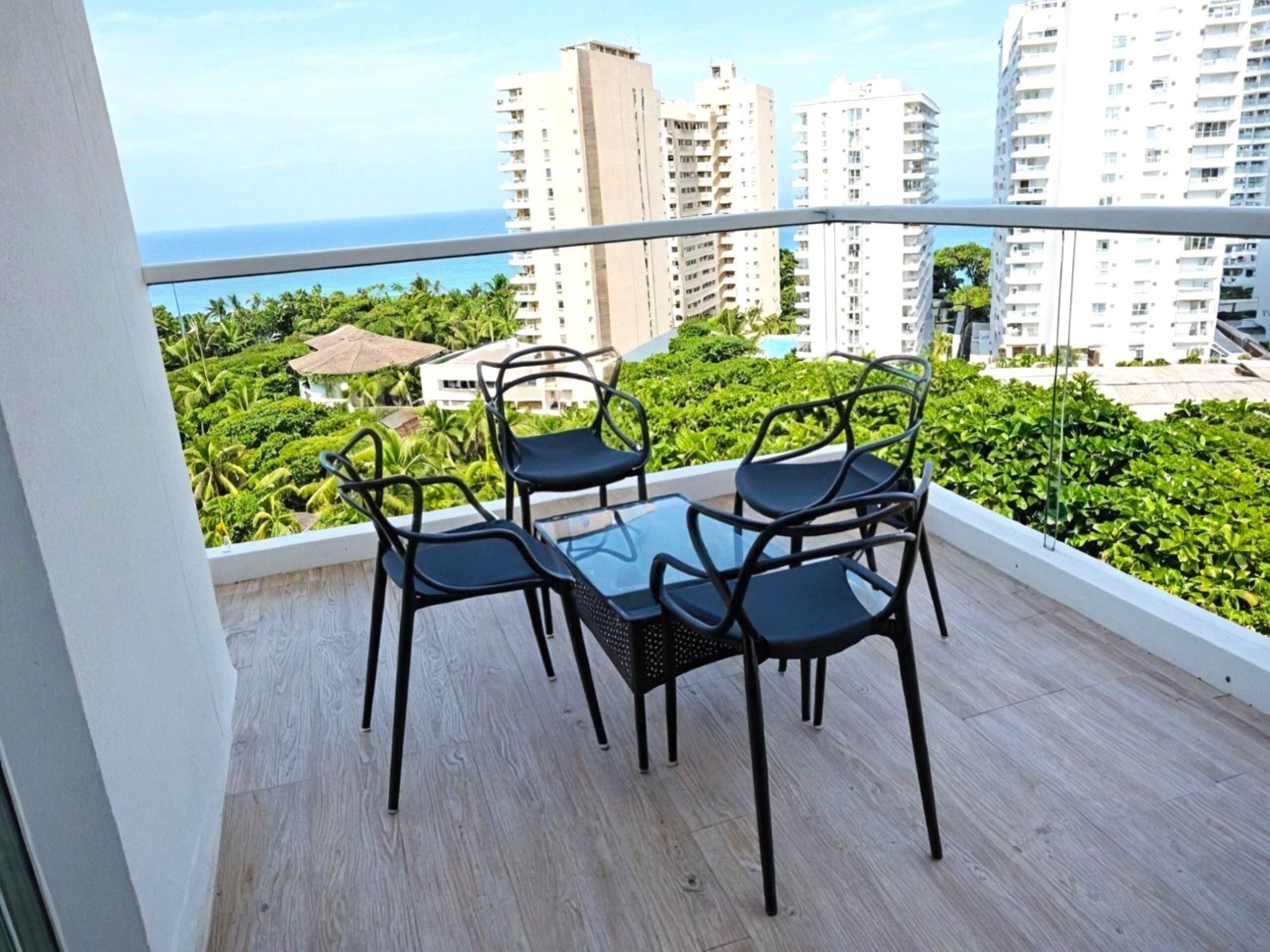 Balcón con una mesa y cuatro sillas, con vista a un paisaje urbano con edificios altos y al océano.