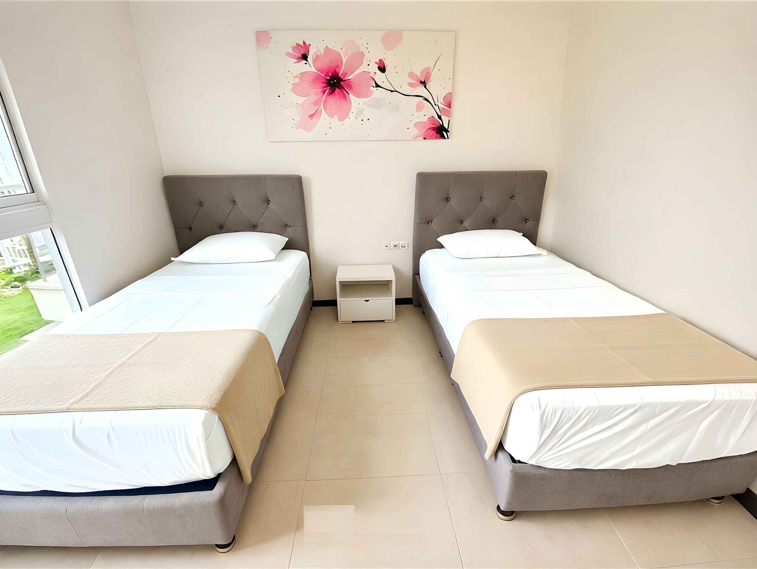 A hotel room with two beds, a small bedside table, a wall-mounted artwork, and a window with a view of greenery.