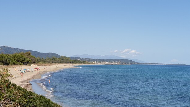 Ein Strand mit klarem, blauem Wasser, einem Sandufer und einem Blick auf eine entfernte Stadtlandschaft mit Gebäuden und Bergen.