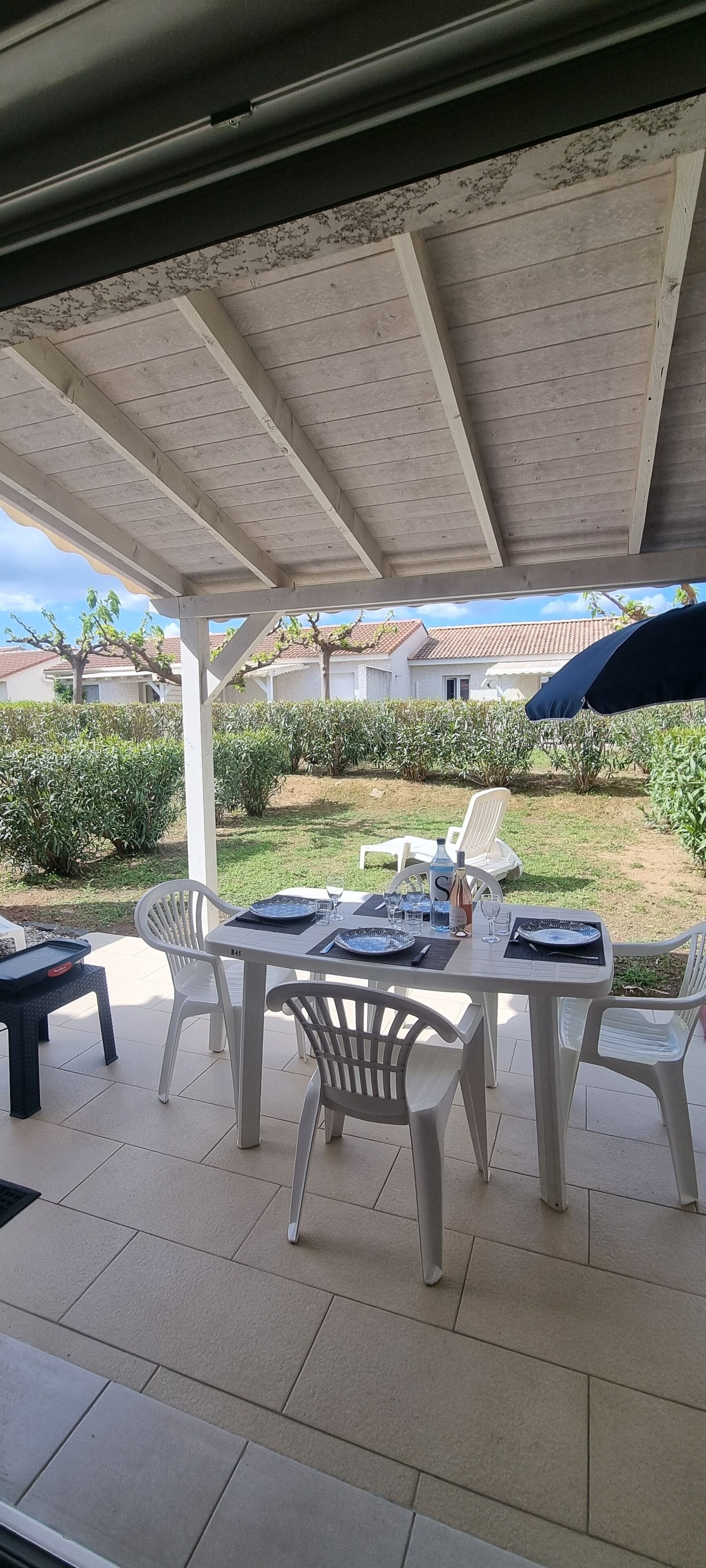 A covered patio with a table set for two, white plastic chairs, and a blue umbrella.