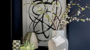 A wooden cabinet with a white vase containing branches and small white flowers, a framed abstract painting, and a checkered object on a tray. - Quiet, convenient & Modern Haven (Gainesville)