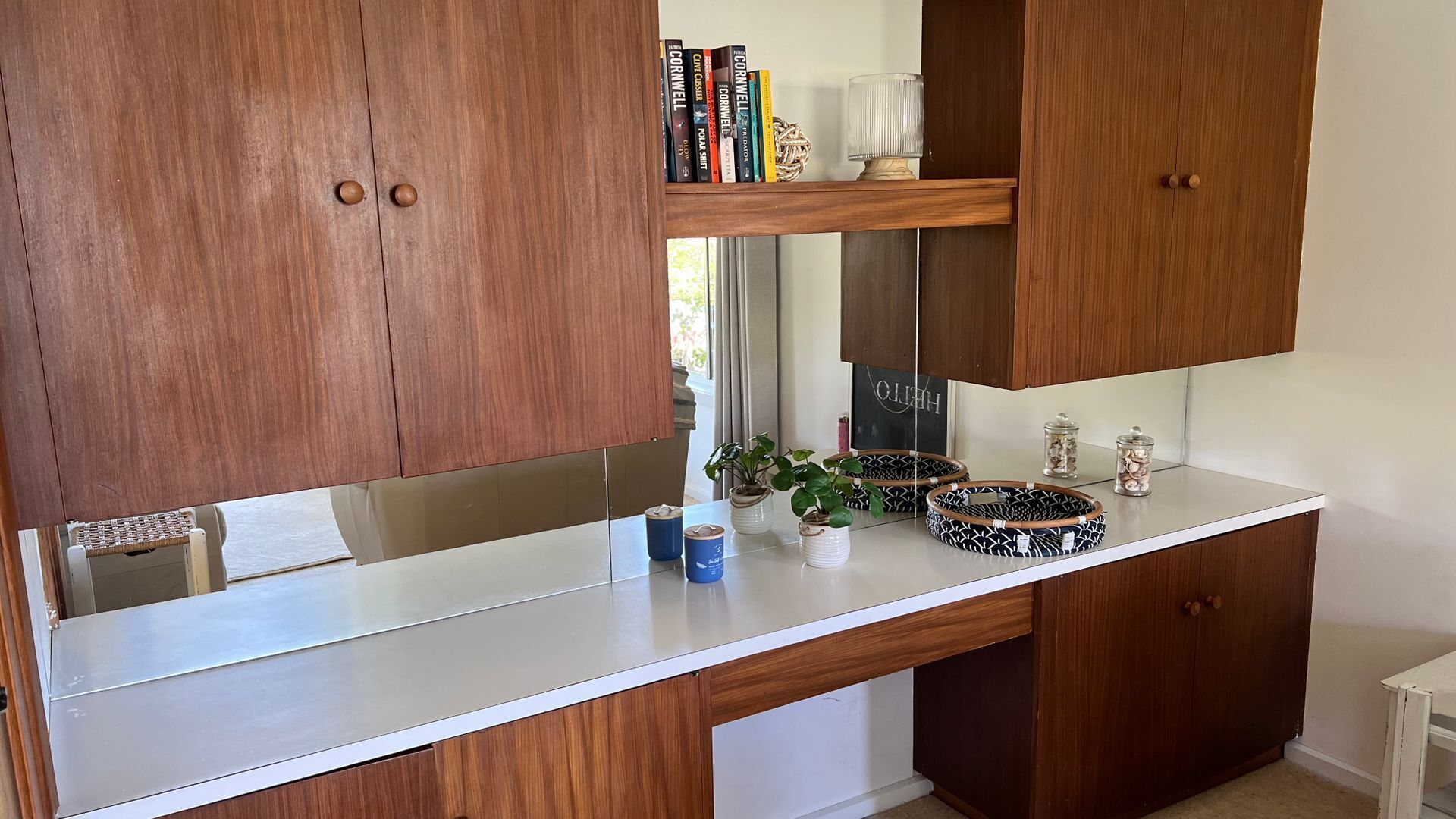A kitchen with wooden cabinets, a white countertop, and a mirror. There are books on a shelf, a plant, and a lamp.
