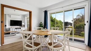 A dining area with a wooden table and white chairs, a TV in the background, and a sliding glass door leading to a garden.