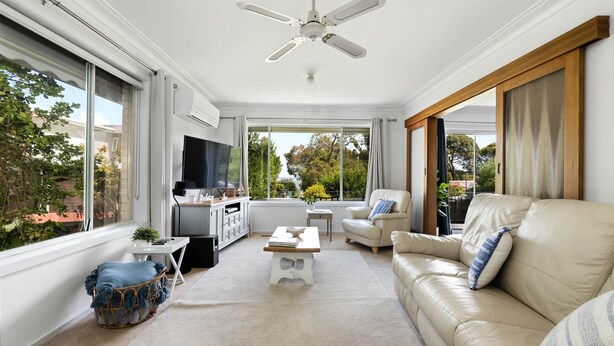 A spacious living room with a large TV, comfortable seating, a ceiling fan, and large windows offering a view of the outdoors.