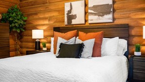 A neatly made bed with a white comforter and multiple pillows in various colors and patterns. There are two framed pictures on the wall above the bed, and a small plant on the nightstand beside the bed. - Peaceful Cabin with Spectacular Stargazing Hot Tub (Alton)