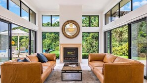A spacious living room with leather sofas, a central fireplace, and large windows offering a view of the outdoors.