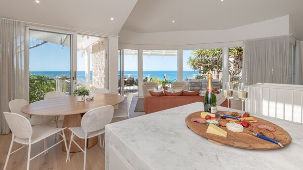 Una habitación amplia con una gran mesa de comedor, sillas de mimbre blancas, una encimera de mármol con una tabla de quesos y vistas al océano.