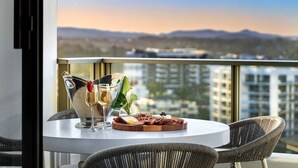 Une table ronde avec un verre de champagne, une bouteille de champagne et une planche de charcuterie garnie de différentes sortes de viandes et de fromages. La table est placée sur un balcon d’où l’on peut admirer un paysage urbain et, au loin, des montagnes.