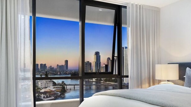 Une chambre d’hôtel avec un grand lit, une table de chevet avec une lampe et une vue sur les toits de la ville, grâce à de grandes fenêtres du sol au plafond.