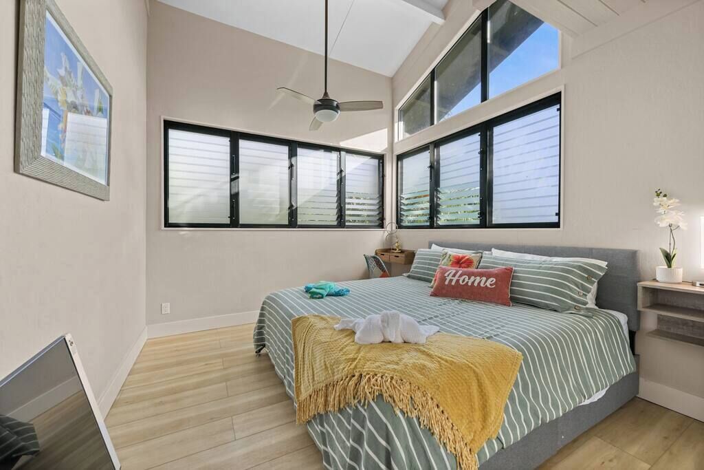 Une chambre à coucher avec un lit, un ventilateur de plafond, de grandes fenêtres et une table de chevet avec une lampe.